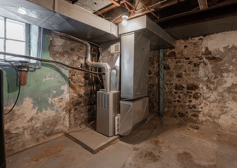 A dimly lit basement with exposed stone walls, featuring a central metal furnace unit connected to ductwork. A small window lets in natural light.