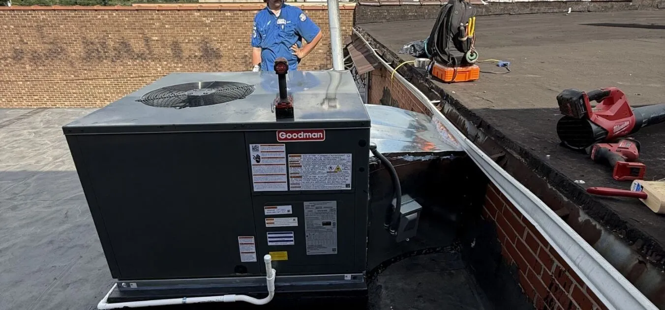Technician servicing a Goodman commercial rooftop air conditioning unit on a flat roof with tools nearby.