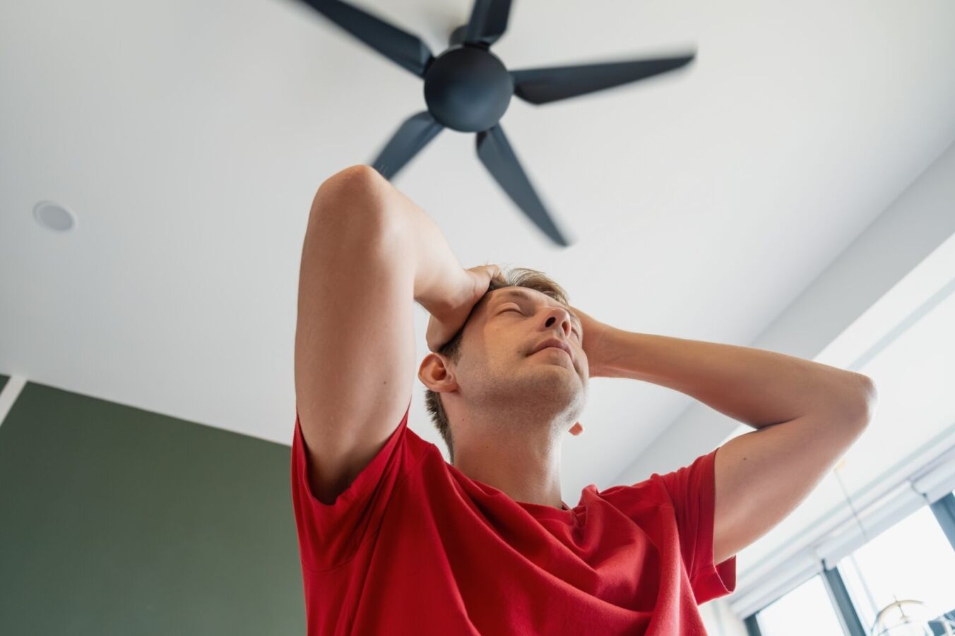 A man sitting under ceiling fan at home, cooling off during hot summer weather, suffering from the high heat.