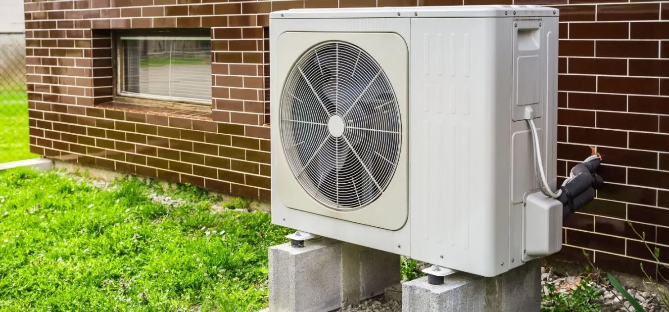 Outdoor heat pump unit mounted on concrete blocks beside a brick house, with green grass and a window visible in the background.