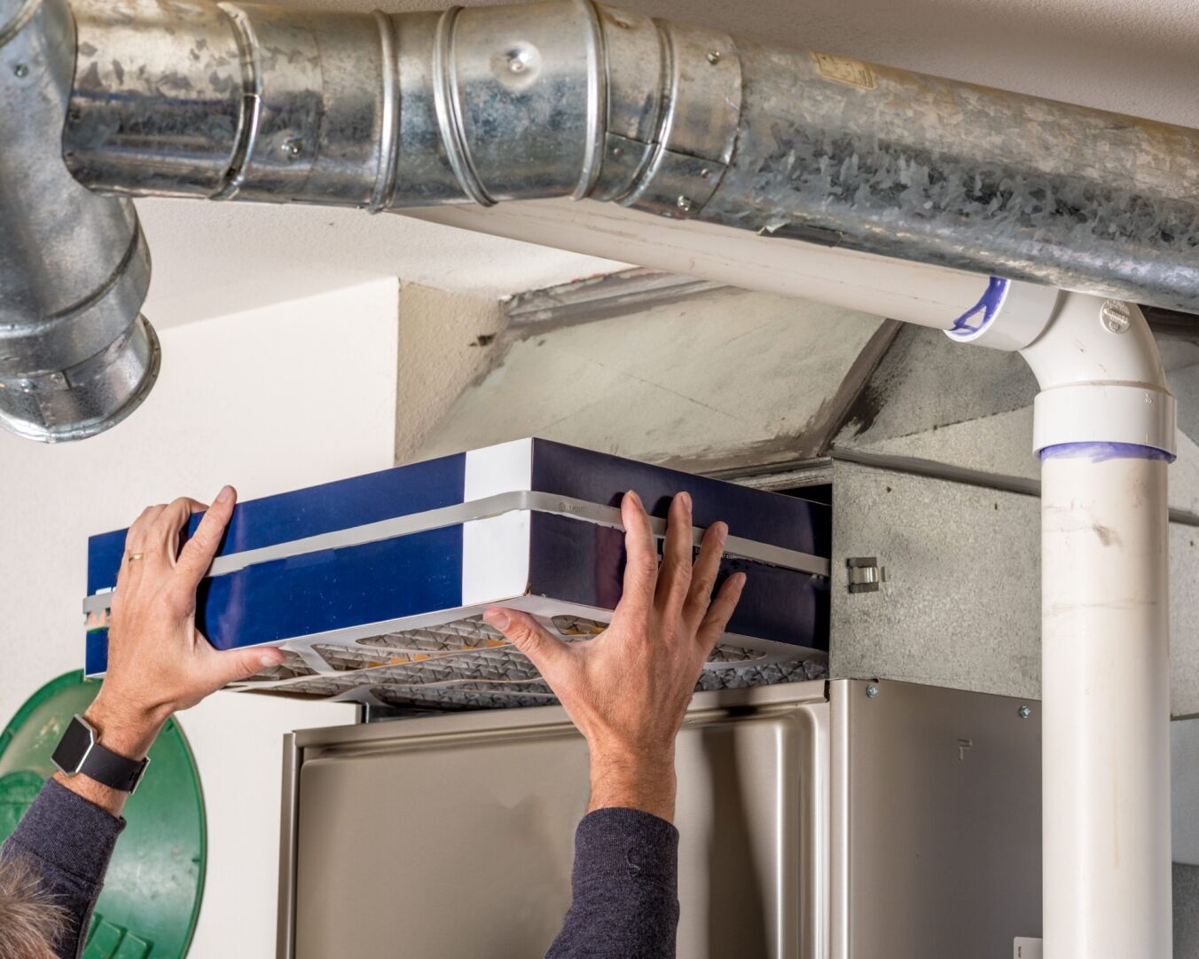 A person is replacing an air filter in an HVAC system, lifting the filter into the unit near exposed ductwork and pipes.