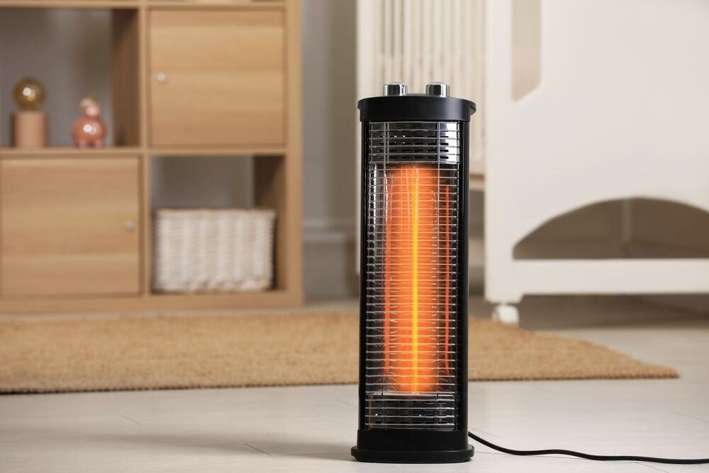 A black vertical space heater with an orange glow is turned on in a room. There is a wooden shelf in the background and a beige rug on the floor.