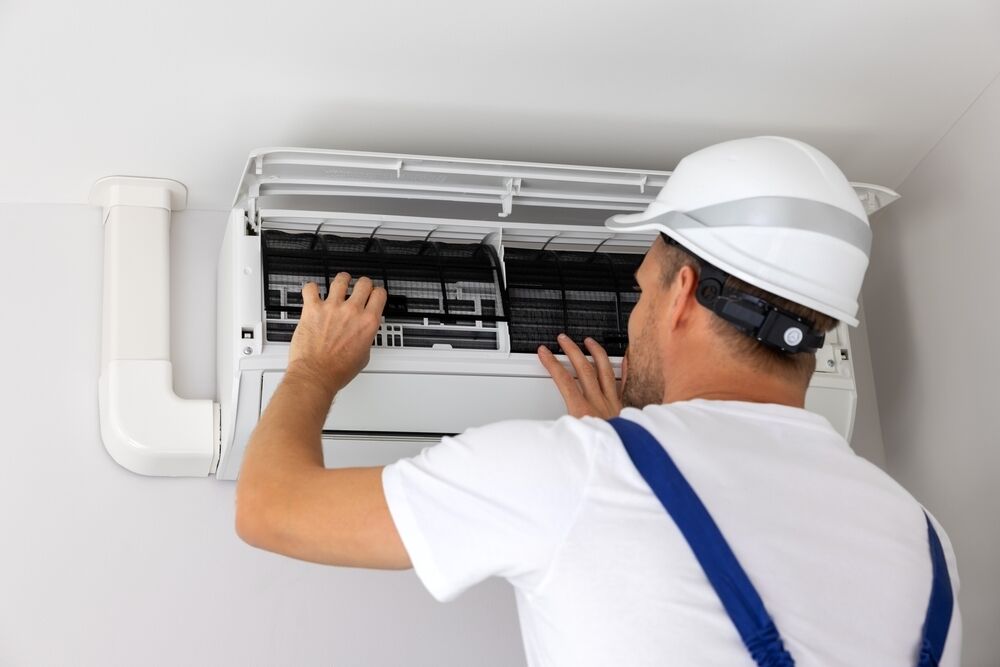A technician in a hard hat and overalls inspects and adjusts an air conditioning unit mounted on a wall.