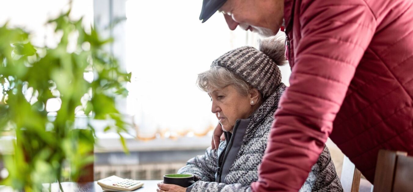 An elderly couple in winter clothing looks at a computer screen together at a table indoors—perhaps searching for advice on what to do when the heat goes out—with a plant and calculator nearby.