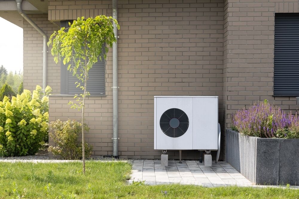 Air conditioning unit installed outside a brick house, next to a small tree and a planter with purple flowers.