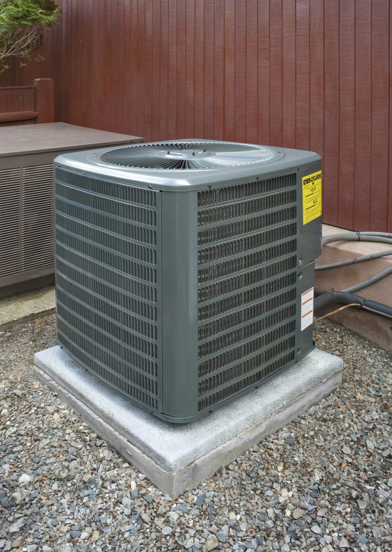 Outdoor central air conditioning unit on a concrete pad with gravel and a wooden fence in the background,