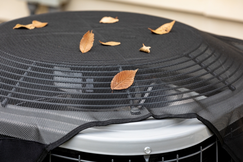 Air conditioning unit outdoors with a protective mesh cover, scattered with dried leaves on top.