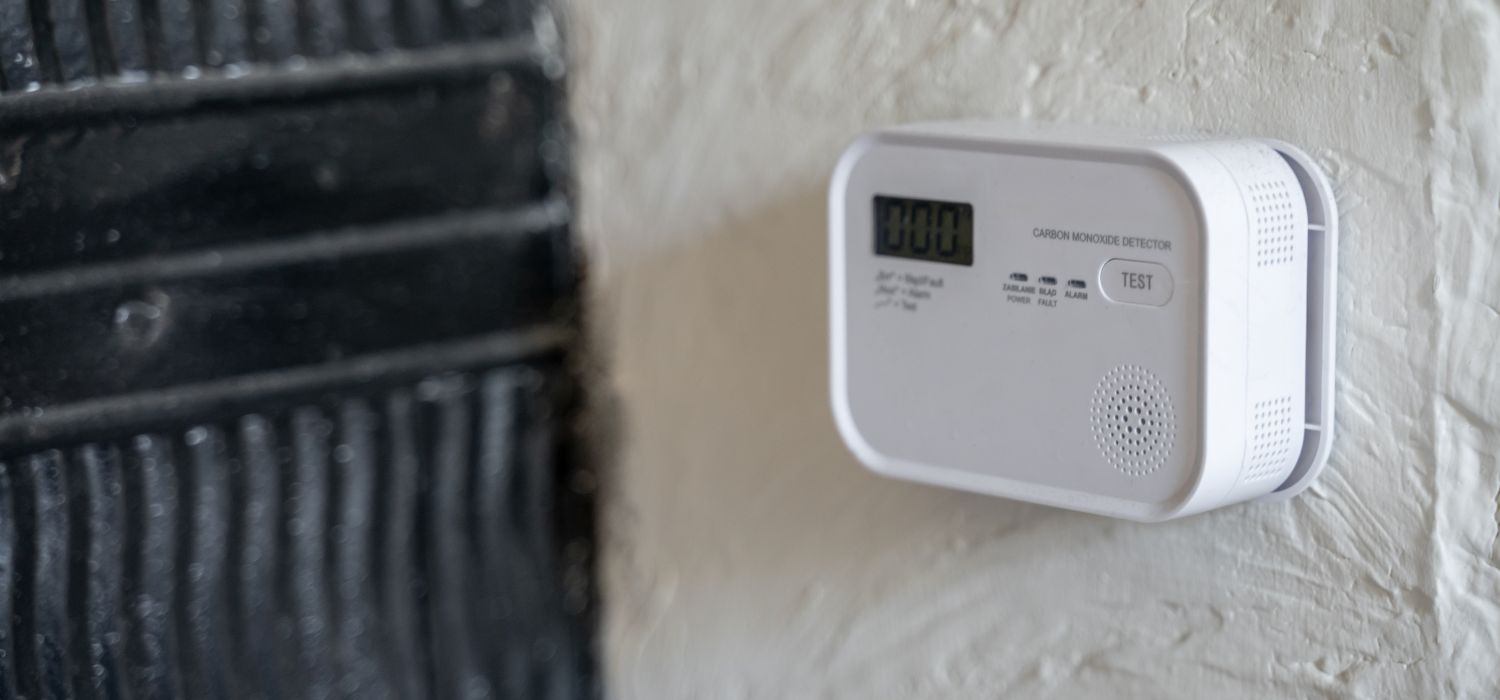 A white carbon monoxide detector with a digital display and test button is mounted on a textured white wall next to a black surface, helping to prevent carbon monoxide in your home.