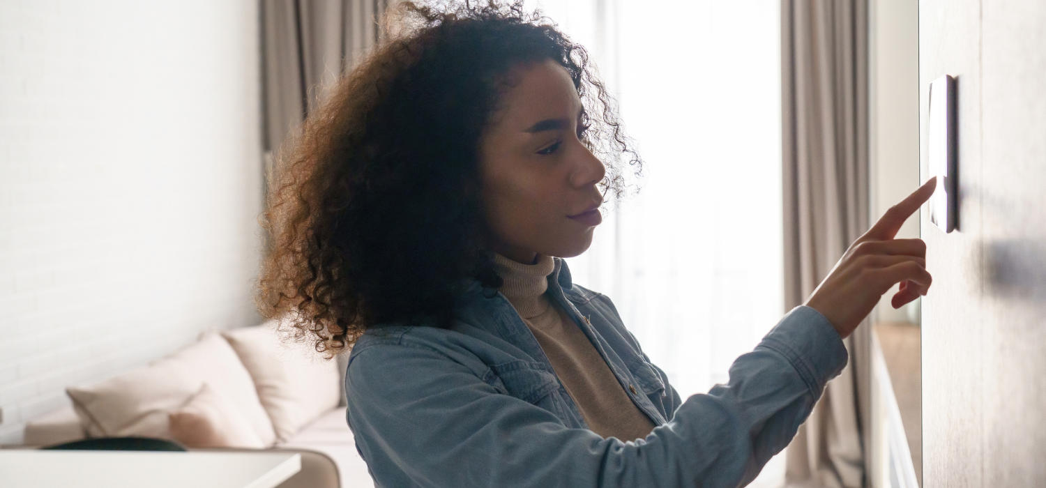 A woman wearing a denim shirt adjusts a smart home thermostat on a wall in a modern living room, setting it to the best winter thermostat settings for comfort and energy efficiency.