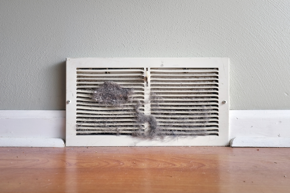 A wall vent covered in dust and lint against a gray wall and wooden floor.