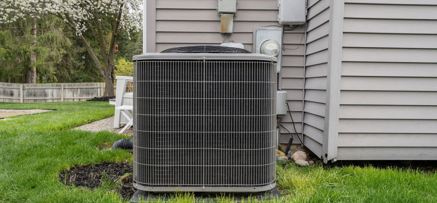Outdoor air conditioning condenser unit installed beside a house on a grassy lawn with a wooden fence and trees in the background.