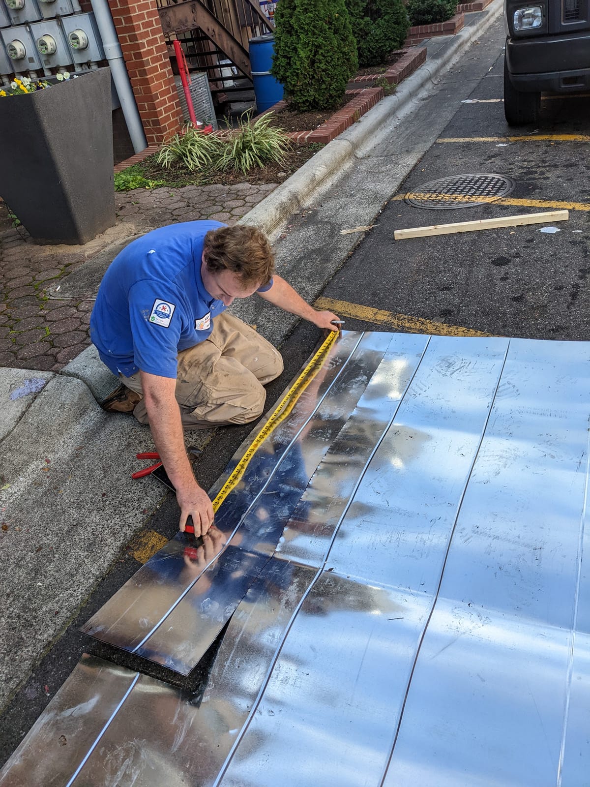 A worker measuring and marking a piece of metal ductwork on the ground for cutting or installation.