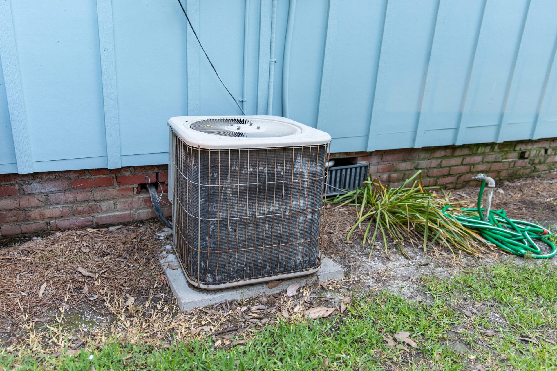An outdoor air conditioning unit beside a house with a garden hose coiled nearby.