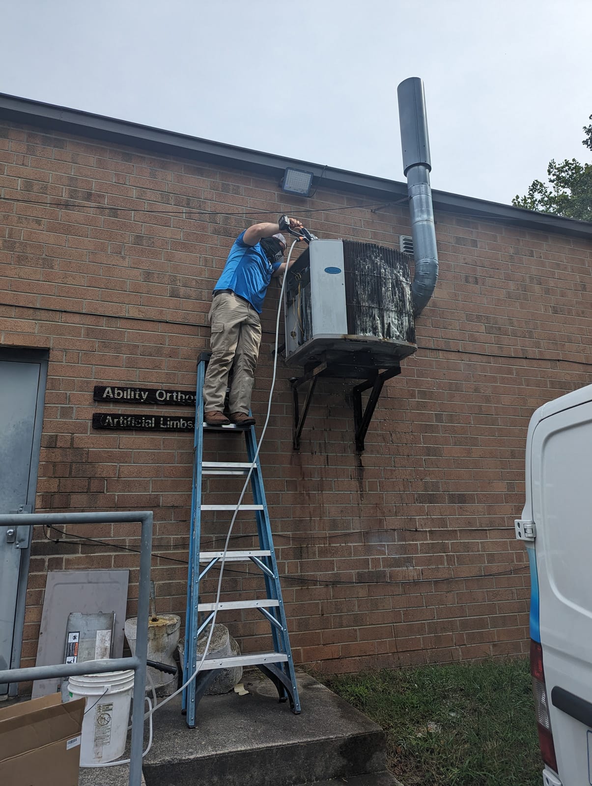A technician on a ladder working on an outdoor hvac unit beside a brick building.