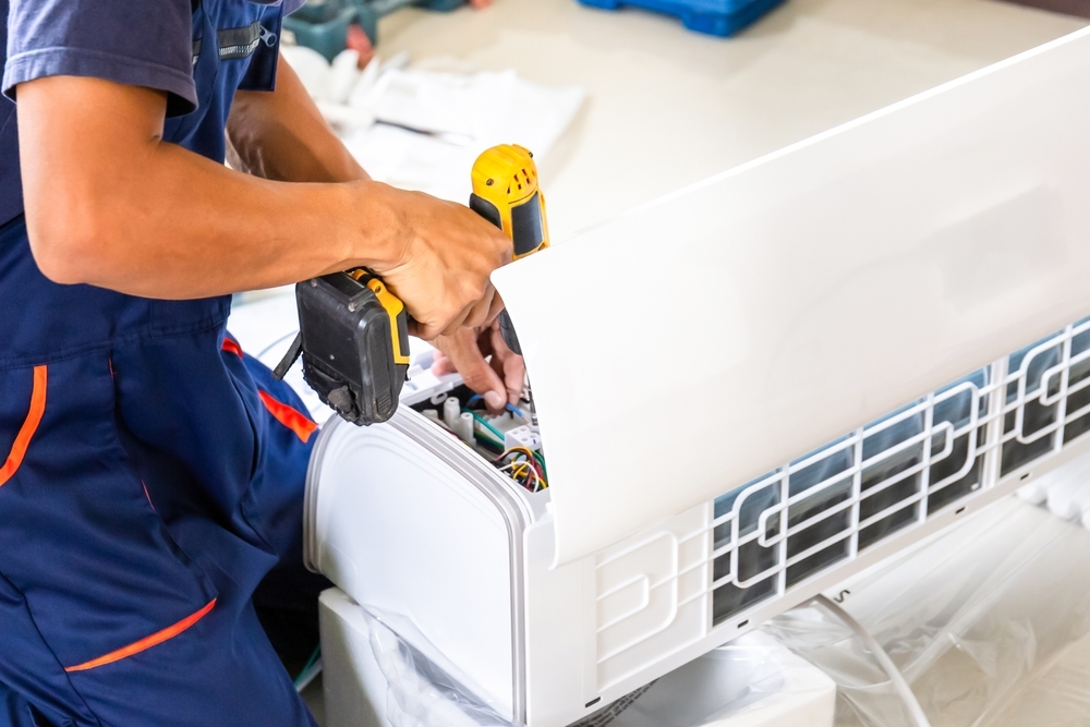 A technician in blue overalls uses a cordless drill to install or repair a wall-mounted air conditioning unit.