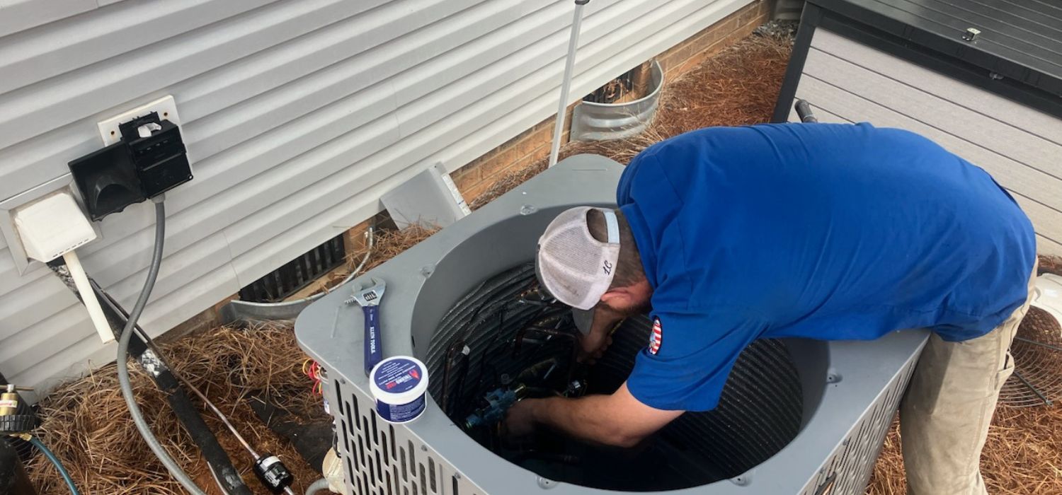 A technician in a blue shirt and white cap is servicing an outdoor air conditioning unit next to a building, performing a Lincolnton NC AC upgrade.