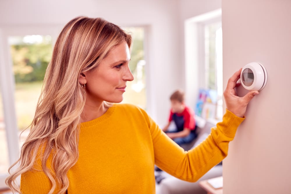 Woman adjusting a thermostat to the ideal HVAC temperature for energy savings on the wall while a child plays in the background.