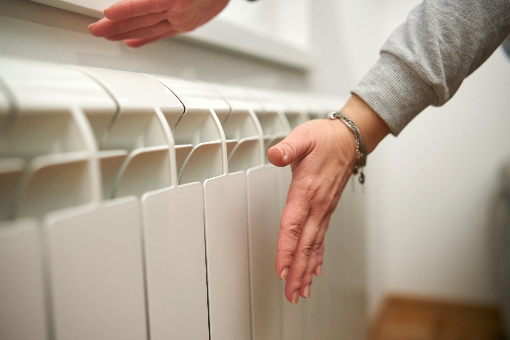 A person holds their hands close to a white radiator, checking for heat, concerned that the furnace is blowing cold air.